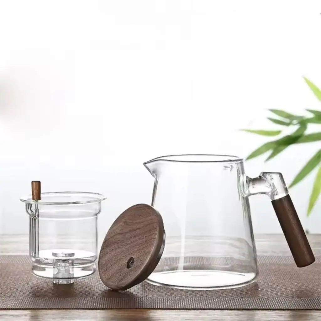 Clear glass pitcher with wooden handle and lid on a light surface with a blurred green plant in the background
