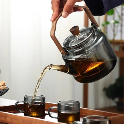 Glass teapot with wooden handle being poured into glass cups on a wooden tray.