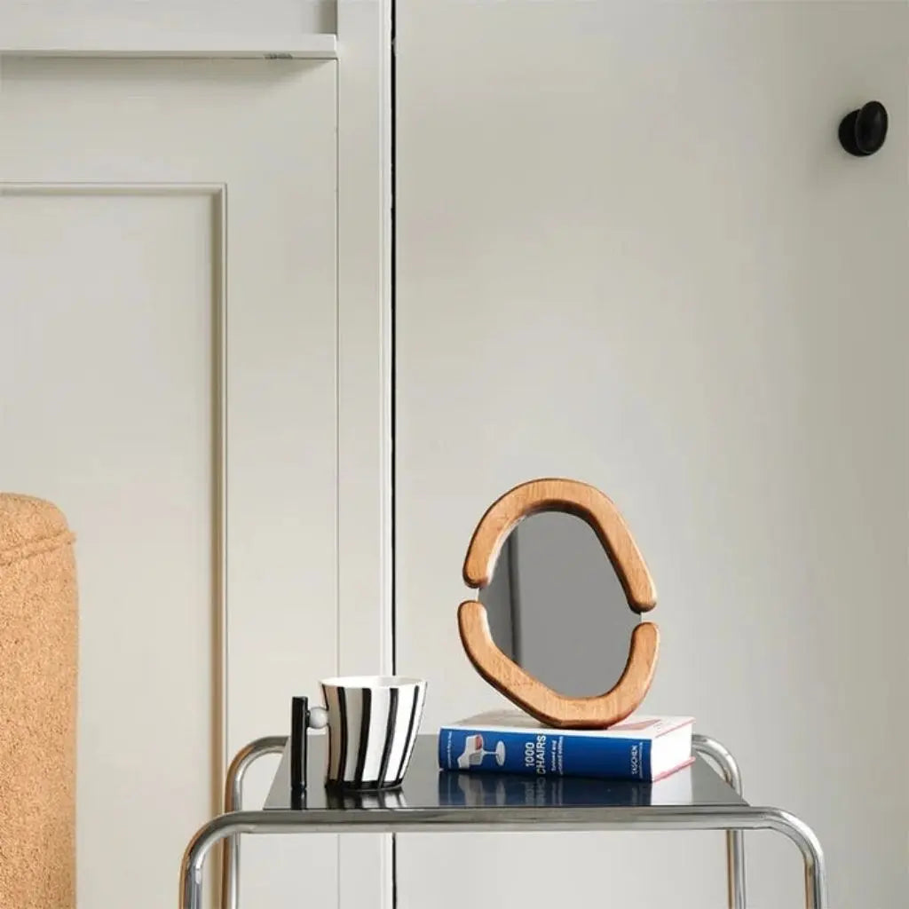 Wooden mirror on a table with books and a cup against a neutral wall.